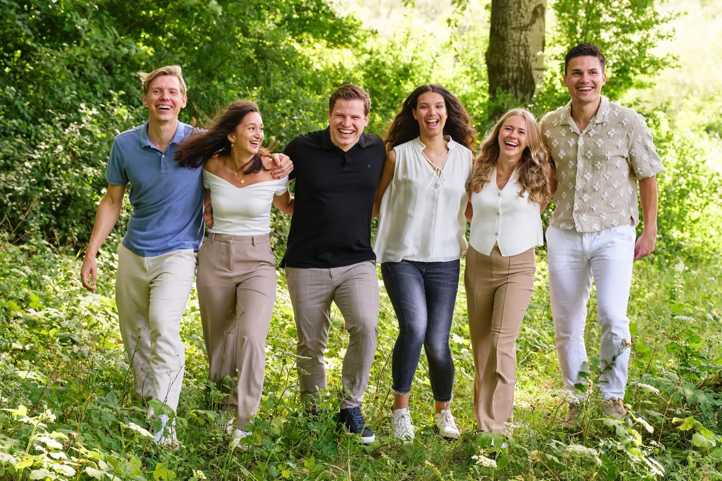 Zes mensen wandelen samen door een groen bos en lachen.