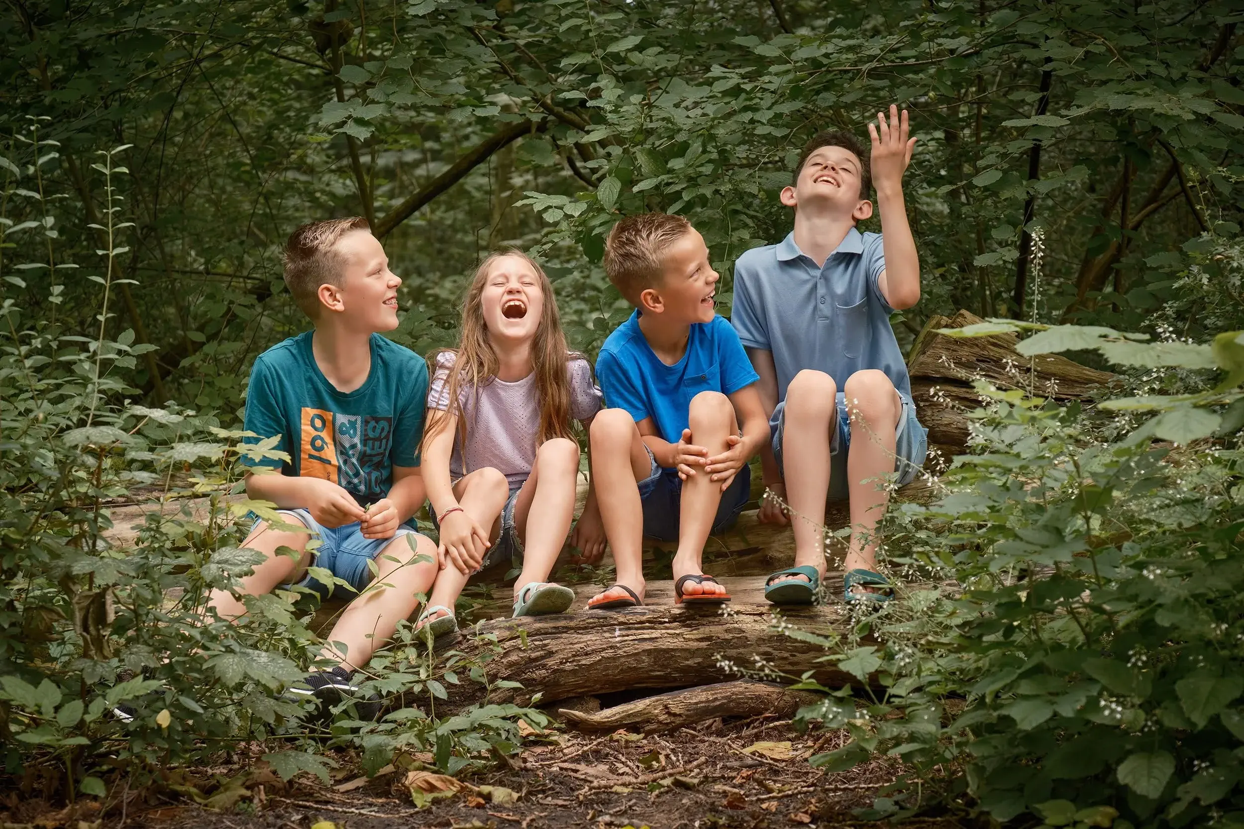 Vijf kinderen zitten op een omgevallen boomstam in een bos, lachend en plezier makend.