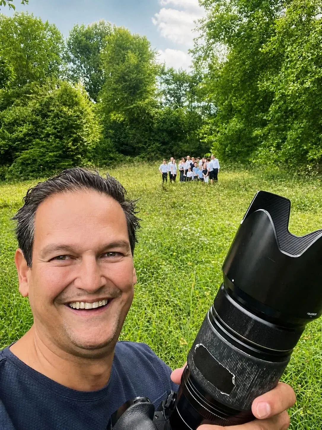 Stefan Segers poseert met zijn camera, met een groep mensen op de achtergrond in het groen