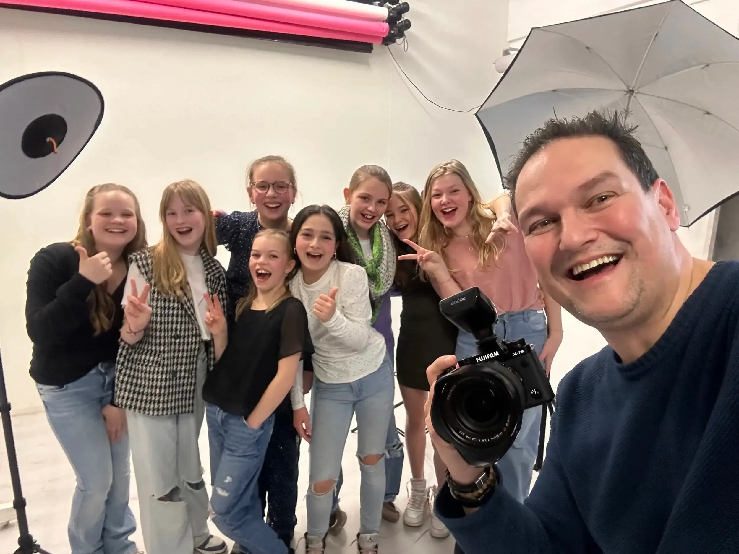 Een groep jonge meisjes poseert voor een foto in een studio, met een man die de camera vasthoudt. De meisjes lachen en maken V-vlaggen met hun handen. De man lacht tevreden aan de rechterkant van de foto.