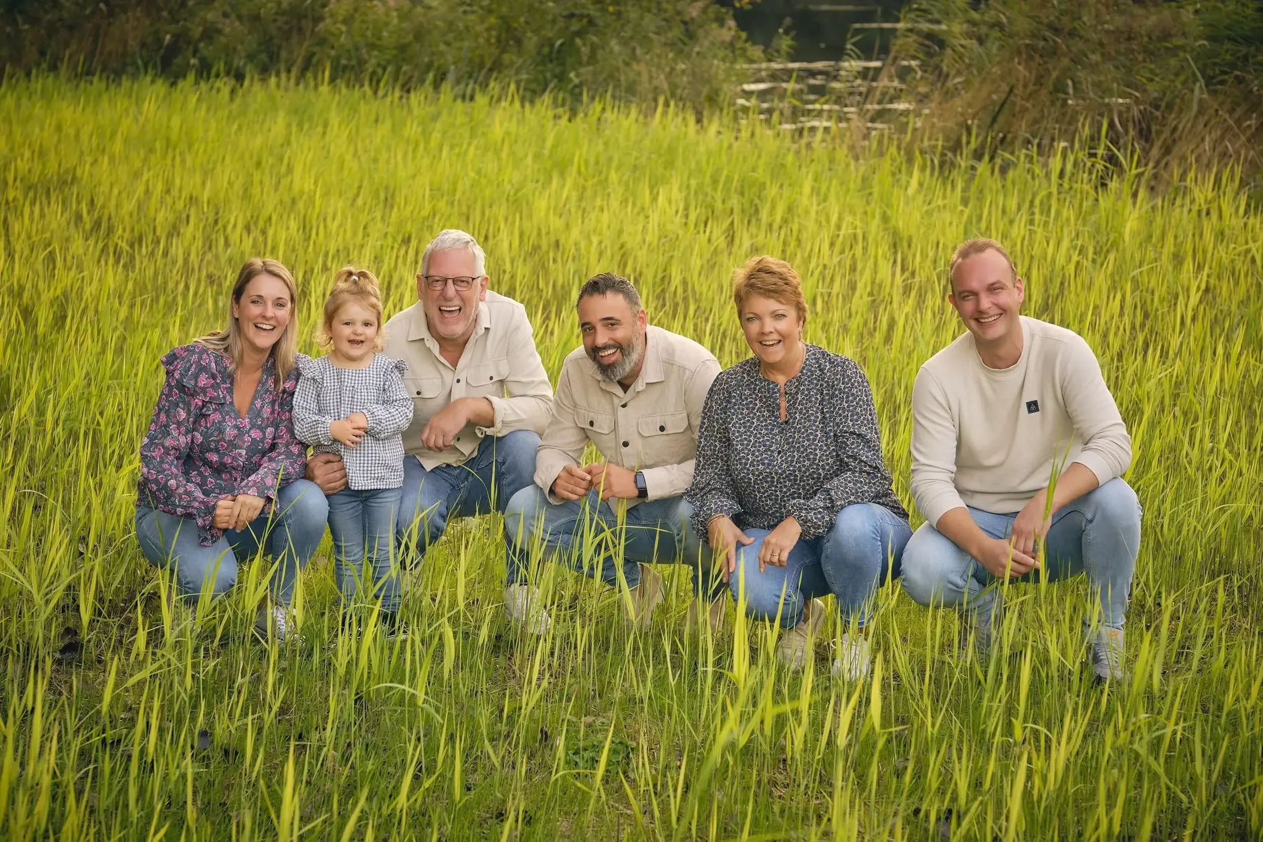 Gezin met vier volwassenen en een kind die glimlachen en poseren in een groene rijstveld buiten, met bomen en water op de achtergrond.