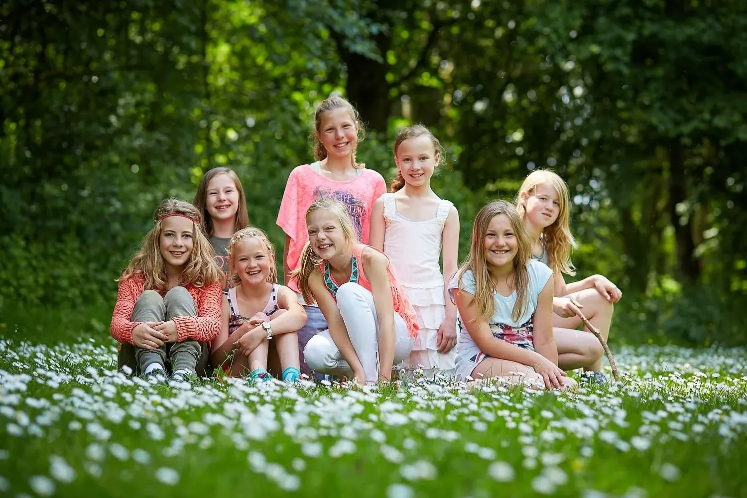 Tien jonge meisjes zitten en staan op een groene, bloemrijke grasweide in het bos, lachend en plezier hebbend.